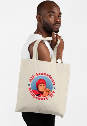 Muž v bílém tílku drží béžovou tašku s potiskem fotbalisty v červené helmě a nápisem "All-American".