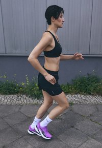 Čierny športový top a šortky, fialové bežecké topánky, biele ponožky s logom. Osoba v polovici kroku, vonkajšie mestské prostredie s betónom a trávou.