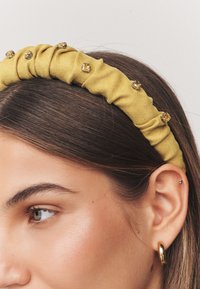 Yellow fabric headband features a twisted design with small decorative gems along one side. Smooth texture and rounded shape.