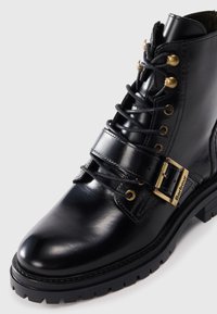 Black leather ankle boot featuring a rounded toe, metal hardware, gold accents, and a thick black rubber sole with a textured grip.