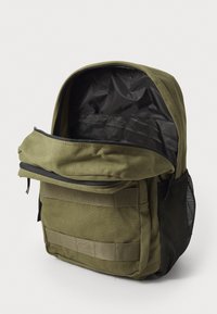 Mochila de lona verde oliva con un compartimento principal con cremallera, bolsillo lateral de malla y correas horizontales en la parte frontal para accesorios.