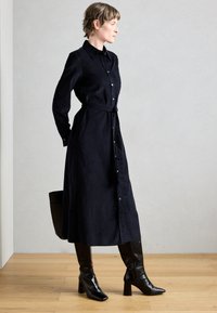 Dlouhé černé košilové šaty s knoflíky, opaskem v pase a dlouhými rukávy, doplněné černými vysokými kozačkami na podpatku ke kolenům.
