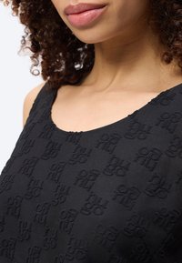 Κοντινό πλάνο ενός ατόμου που φοράει μαύρο, υφασμάτινο αμάνικο τοπ με επαναλαμβανόμενα ανάγλυφα αφηρημένα σχέδια. Μερικώς ορατά σγουρά μαλλιά.