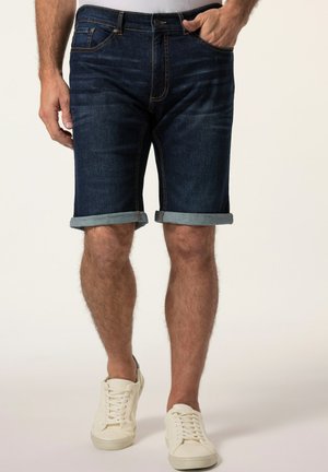 Mand iført mørkeblå optrukne denimshorts og hvide sneakers med afslappede hænder ned langs siderne.