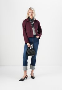 Veste en laine marron avec poches avant, blouse grise, jeans bleus avec revers, chaussures à talons slingback noires et sac à main noir texturé.
