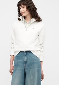 Femme aux cheveux bouclés portant un sweat-shirt blanc à fermeture éclair partielle sur une chemise à col et un jean bleu taille haute à jambes larges, mains détendues.