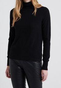 Pull à col roulé noir en maille douce avec manches longues, coupe ajustée et poignets côtelés. Porté avec un pantalon en cuir noir.