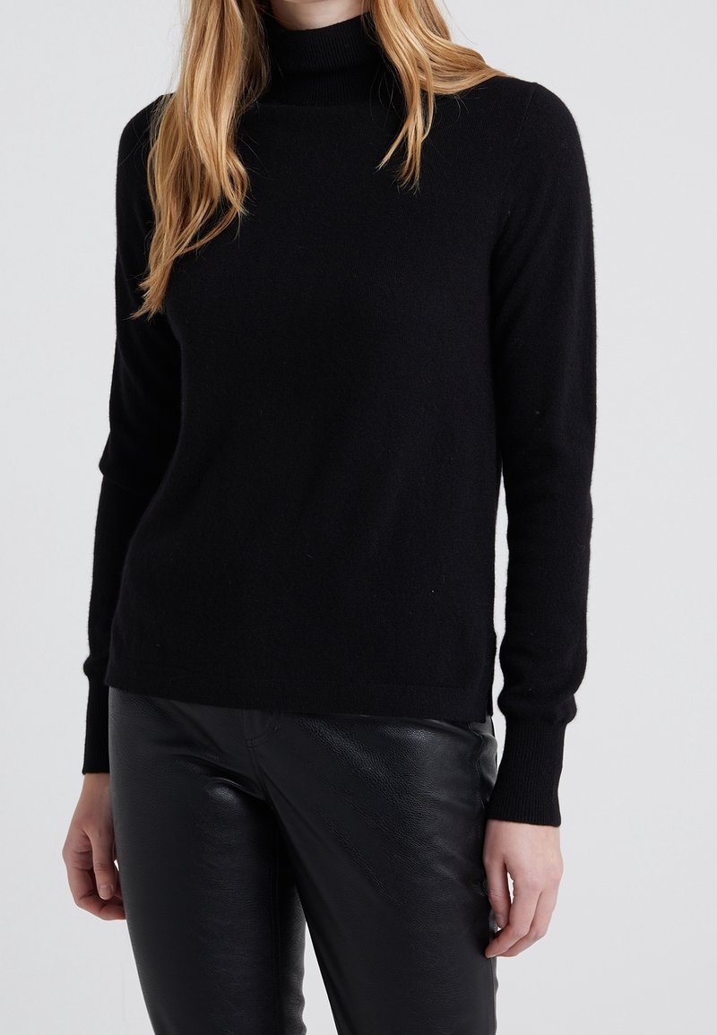 Pull à col roulé noir en maille douce avec manches longues, coupe ajustée et poignets côtelés. Porté avec un pantalon en cuir noir.