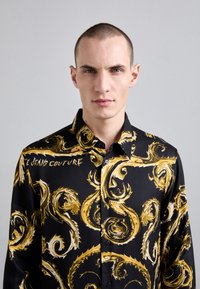 Maglietta nera con motivi dorati a spirale, con design a bottoni e colletto a punta. Testo "JEANS COUTURE" stampato sul petto.