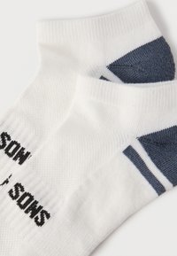 Paire de chaussettes blanches à la cheville avec des accents gris foncé au talon et aux orteils, et un texte noir sur la semelle, posées sur un fond blanc.