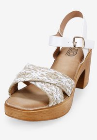 Sandales à talons en cuir blanc avec des brides en fibres naturelles tressées, un talon en bois, et une sangle de cheville réglable avec une boucle dorée.