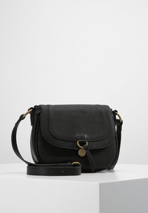 Zwarte leren crossbodytas met klepsluiting, verstelbare band en kleine ronde gouden hanger op een wit displayplateau.