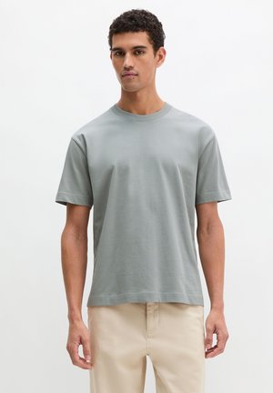 Junger Mann trägt ein hellgraues kurzärmeliges T-Shirt und beige Hosen und steht vor einem schlichten weißen Hintergrund.