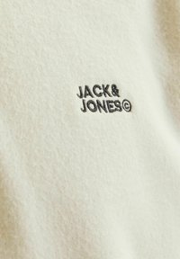 Krämmfärgad textil med en sammetsliknande textur och en svart broderad logotyp med texten "JACK & JONES." En enkel design och mjuk yta.