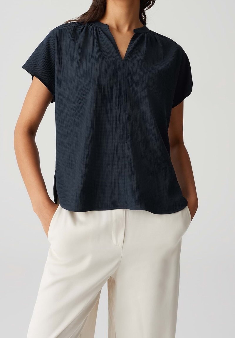 Haut bleu marine à manches courtes style cache-cœur avec un motif texturé et des fentes latérales, associé à un pantalon crème.