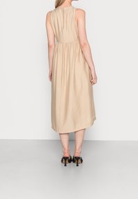 Beige bezramenné šaty s přiléhavým pasem a volnou sukní, které mají jemnou strukturu a zipovou zadní část. Nohy doplněné černými podpatky.