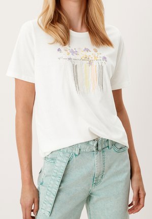 Vrouw draagt een wit T-shirt met geborduurde wilde bloemen en tekst, gecombineerd met lichtblauwe hooggetailleerde jeans met een stoffen riem.