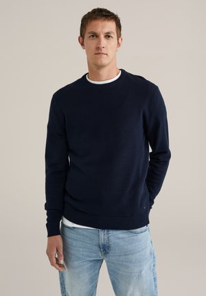 Hombre con un suéter de punto azul marino sobre una camiseta blanca y jeans de mezclilla claros, de pie con una mano en el bolsillo frente a un fondo liso.