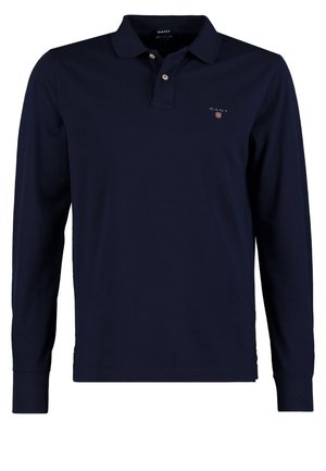 Polo à manches longues bleu marine, jean skinny noir et baskets bordeaux. Sac à bandoulière marron avec des accents en cuir à la main.