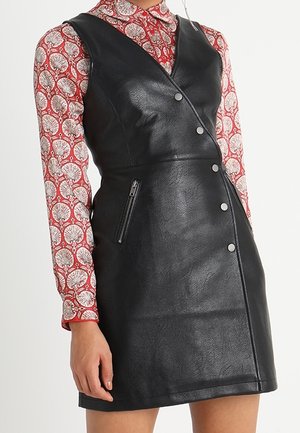 Vrouw draagt een zwarte mouwloze V-halsjurk van kunstleer met knopen aan de voorkant en een ritszak, over een patroonrijke blouse met lange mouwen.