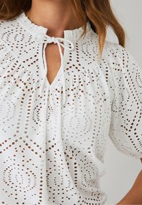 Femme portant un blouse en dentelle blanche à œillets avec des manches bouffantes et un décolleté en trou de serrure noué, révélant des motifs découpés circulaires complexes.