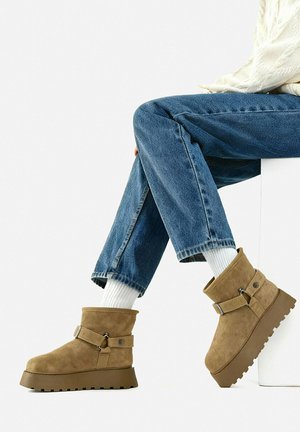 Persona con jeans blu e calzini bianchi seduta su un blocco bianco, indossando stivaletti con zeppa in camoscio color tan con cinturini.