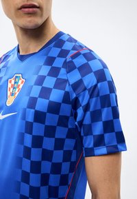 Κοντινό πλάνο ατόμου που φοράει μπλε αθλητική φανέλα με καρό σχέδιο, με το έμβλημα της HNS και λευκό λογότυπο Nike στο στήθος, μπροστά σε απλό φόντο.