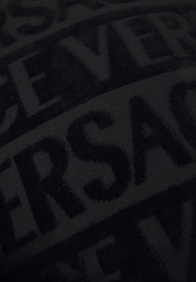 Juodas audinys su iškilniu tekstu "Versace." Tekstūra yra minkšta, atskleidžianti matinės ir blizgios apdailos kontrastą rašte.
