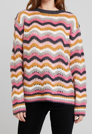 Femme portant un pull en maille manches longues avec des rayures ondulées multicolores en rose, blanc, moutarde, gris et noir, associé à un pantalon noir.