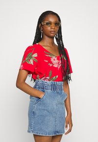 Blusa curta de manga comprida com padrão floral vermelho, com padrões verdes e rosas, combinada com uma saia de ganga azul clara com cintura elástica e bolsos.