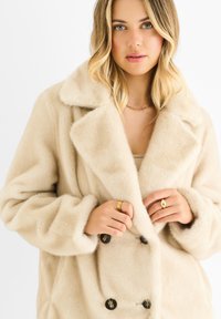 Beige double-breasted coat made of soft faux fur with large notched collar and dark buttons. Features wide cuffs and a relaxed fit.