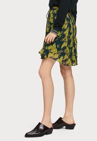 Jupe plissée à motif floral dans un vert foncé avec des fleurs jaunes, dotée d'un ourlet volanté et d'une taille élastique. Des mules noires à talons complètent le look.