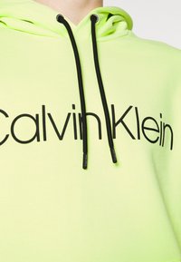 Felpa con cappuccio verde lime con cordini neri e logo "Calvin Klein" nero prominente sul davanti. Realizzata in tessuto morbido.