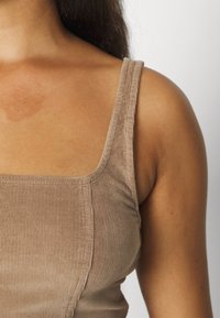 Lysebrun corduroy-top med brede stropper og firkantet halsudskæring, der fremhæver tekstur og ribbet mønster. Nærbillede af den øverste del af overkroppen.