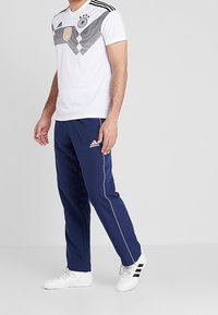 Man klädd i en vit Tyskland fotbollsjersey med svarta ränder och marinblå Adidas träningsbyxor med vit piping samt vita Adidas sneakers.