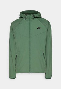Grüne Nike-Jacke mit einem Reißverschluss an der Vorderseite, Kapuze und zwei Seitentaschen. Verfügt über schwarze Akzente und ein Logo auf der Brust. Leichtes Material.