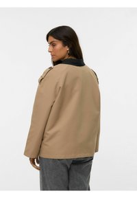 Beige vandtæt jakke med en sort krave, minimalistisk design og skulderknapper. Stoffet ser glat og mat ud i tekstur.