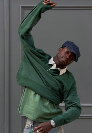 Mees, kes kannab tumerohelist särki ja mereväe sinist nokamütsi, sirutab end ühe käega, mis on tõstetud halli paneelseina taustal.