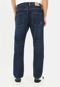 Dunkelblaue Jeans aus Denim mit geradem Bein, Gesäßtaschen und einem hellbraunen Lederpatch am Bund. Tragen mit weißen und marineblauen Sneakers.
