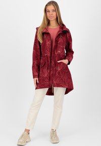 Giacca a vento bordeaux con stampa di foglie, due tasche frontali, chiusura con zip, vita con cordino e cappuccio. Abbinata a pantaloni chiari e sneakers.