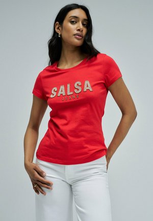 Mujer con camiseta roja ajustada con el texto "SALSA JEANS" y pantalones blancos, posando con una mano en la cadera frente a un fondo liso.