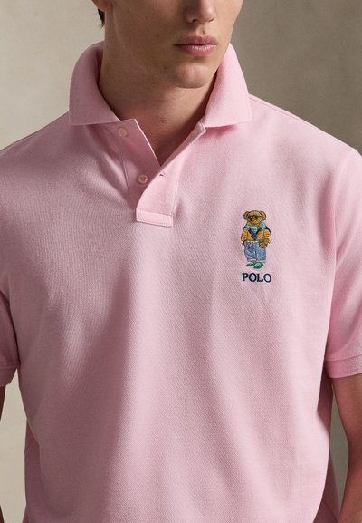 Man wearing light pink polo shirt with embroidered bear in casual clothes and the word "POLO" below on left chest.