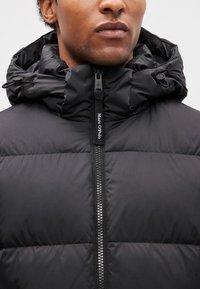 Veste matelassée noire avec fermeture éclair à l'avant, dotée d'un col montant et d'une capuche ajustable. Texture douce et matelassée, tirette zippée à l'effigie de la marque.