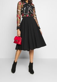 Donna che indossa una blusa trasparente nera a fiori con fiocco, una gonna midi nera e stivaletti neri, mentre tiene una piccola borsa rossa su uno sfondo bianco.