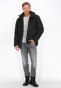 Sort polstret jakke med hætte, der har lynlås og trykknaplukninger, parret med en lys grå sweatshirt og grå jeans. Sorte støvler fuldender outfittet.