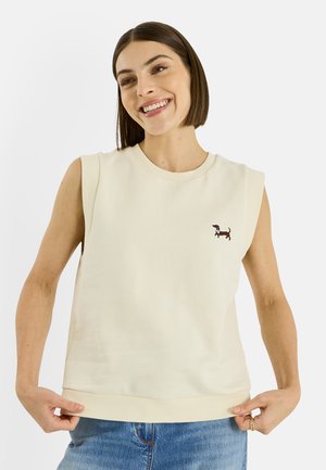 Mujer sonriente con cabello castaño liso que lleva una camiseta sin mangas color crema con un pequeño logo de dachshund y jeans azules, sosteniendo el borde de la camiseta.