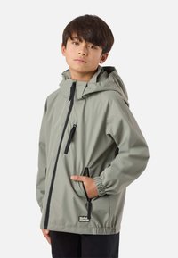 Ragazzo con capelli castano scuro e lisci, indossa una giacca con cappuccio grigio chiaro con dettagli delle cerniere nere, in piedi con una mano in tasca contro uno sfondo bianco.