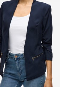 Blazer bleu marine à coupe ajustée, tissu léger et poches latérales zippées. Porté sur un haut blanc et associé à un jean taille haute.