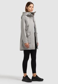 Femme portant un imperméable à capuche gris clair, des leggings noirs et des baskets noires, debout devant un fond uni.