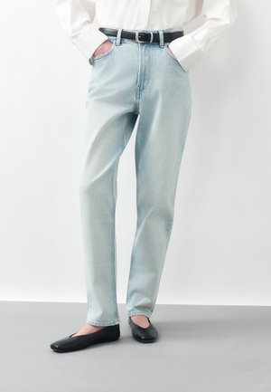 Osoba ubrana w jasnoniebieskie proste jeansy, czarny pasek, białą koszulę z długim rękawem, ręce w kieszeniach i czarne płaskie buty stojąca na szarej podłodze.